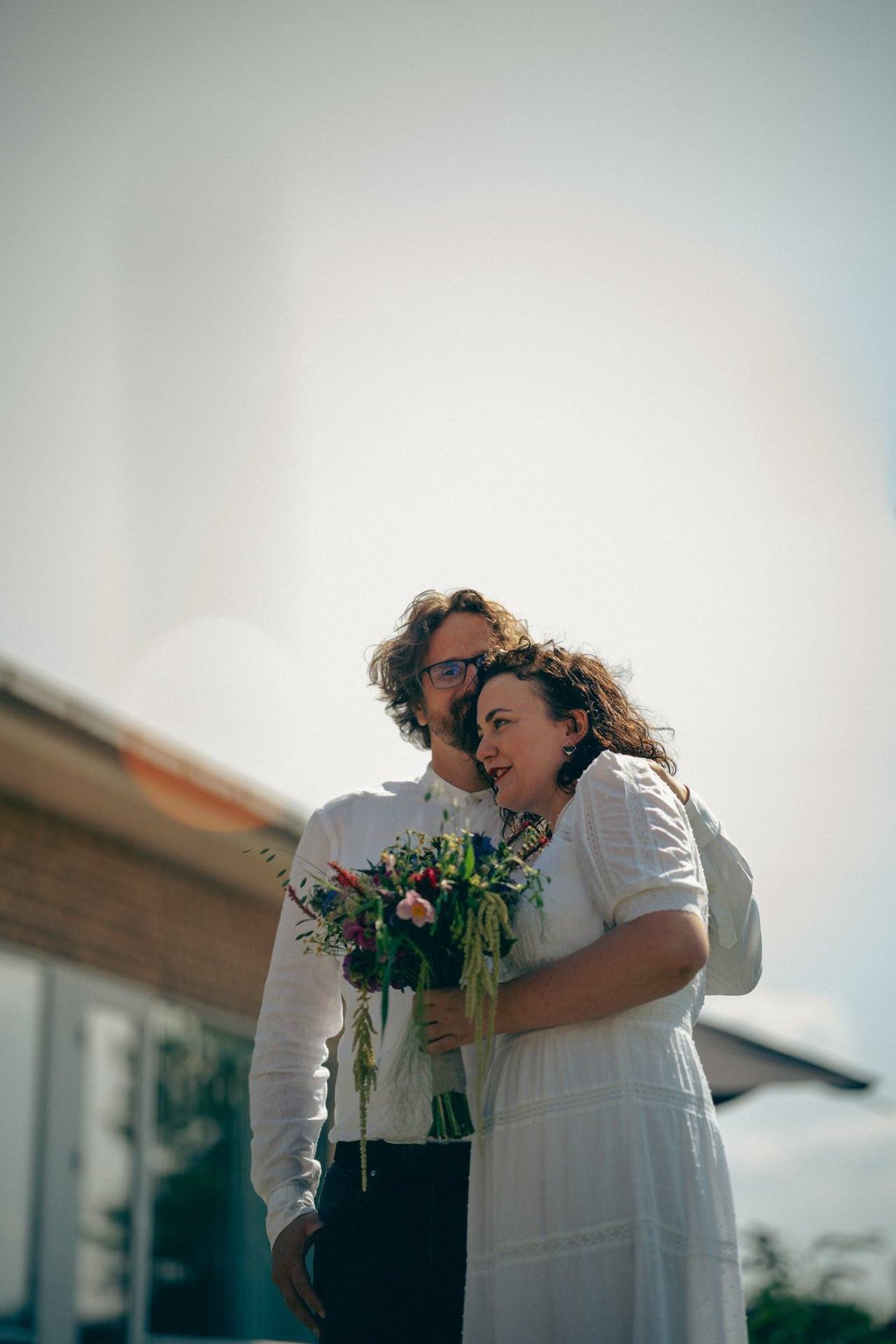 Ein Paar bei einer Hochzeit, das im Freien steht, die Frau hält einen bunten Blumenstrauß, beide lächeln und schauen sich glücklich an.