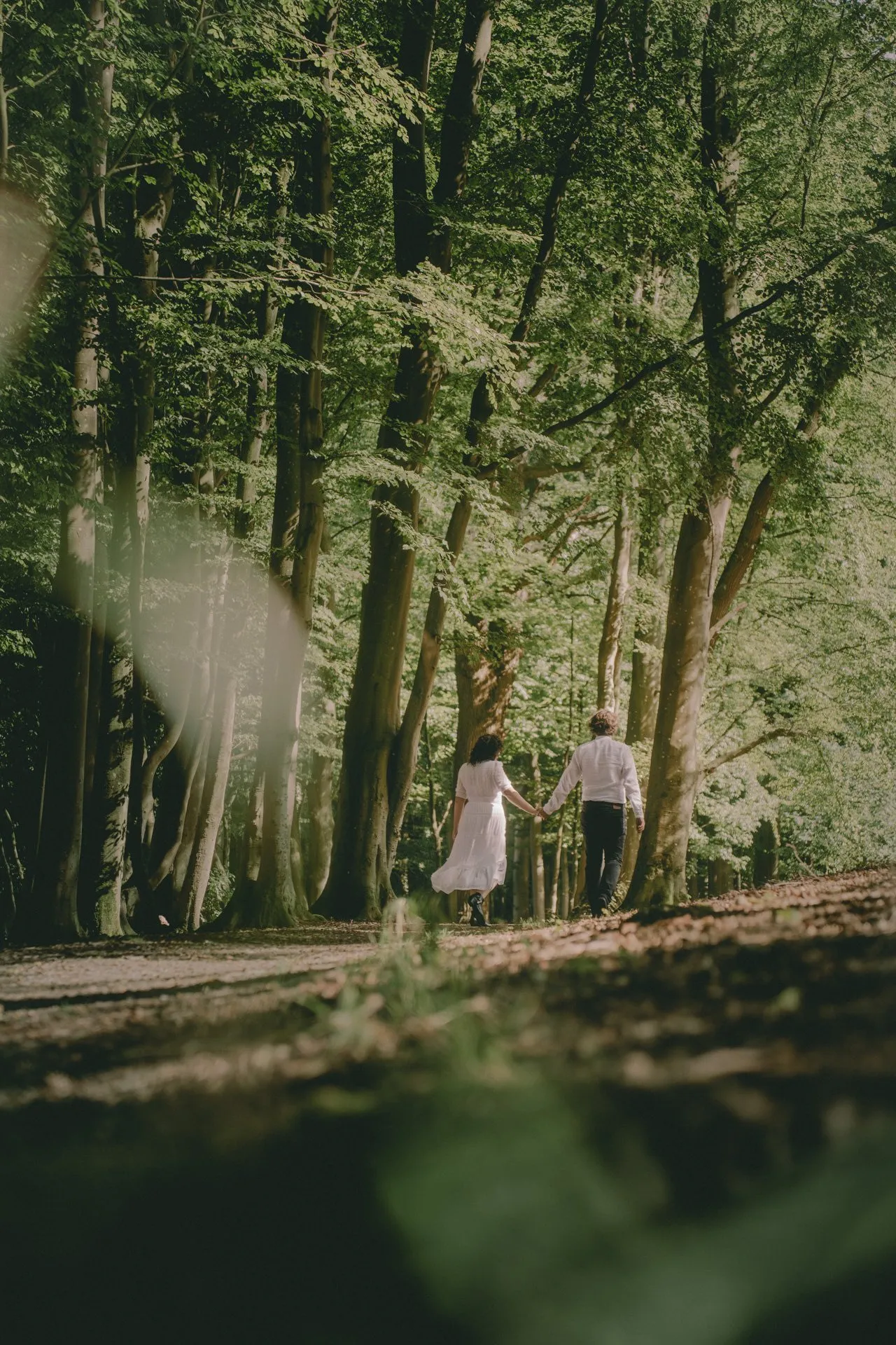Ein Paar hält sich an einem Walddamm die Hand und geht durch einen grünen Wald, die Frau trägt ein weißes Kleid, der Mann trägt helle Kleidung.