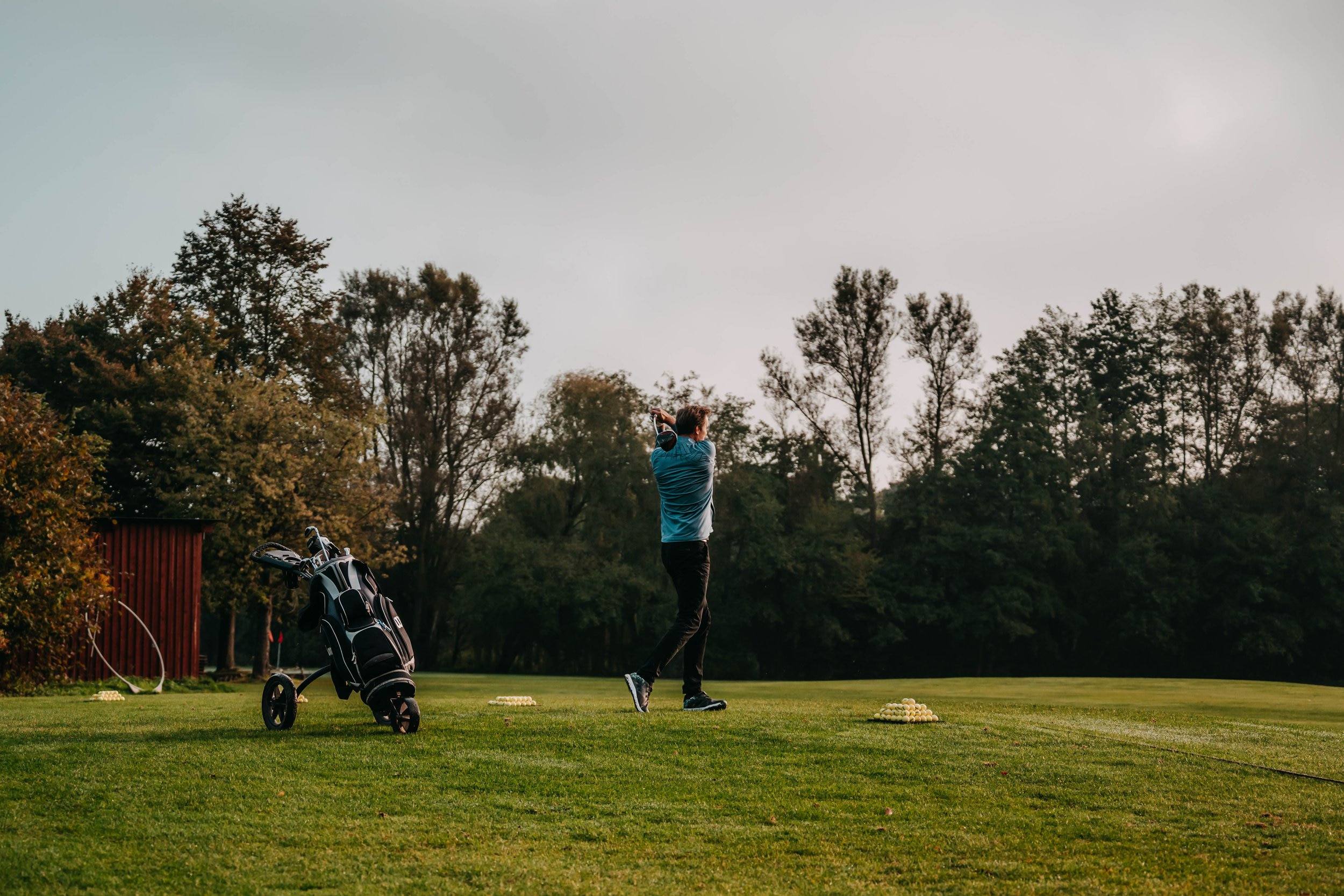 Golfer beim Abschlag auf der Driving Range, daneben ein Golftrolley, Bäume im Hintergrund.