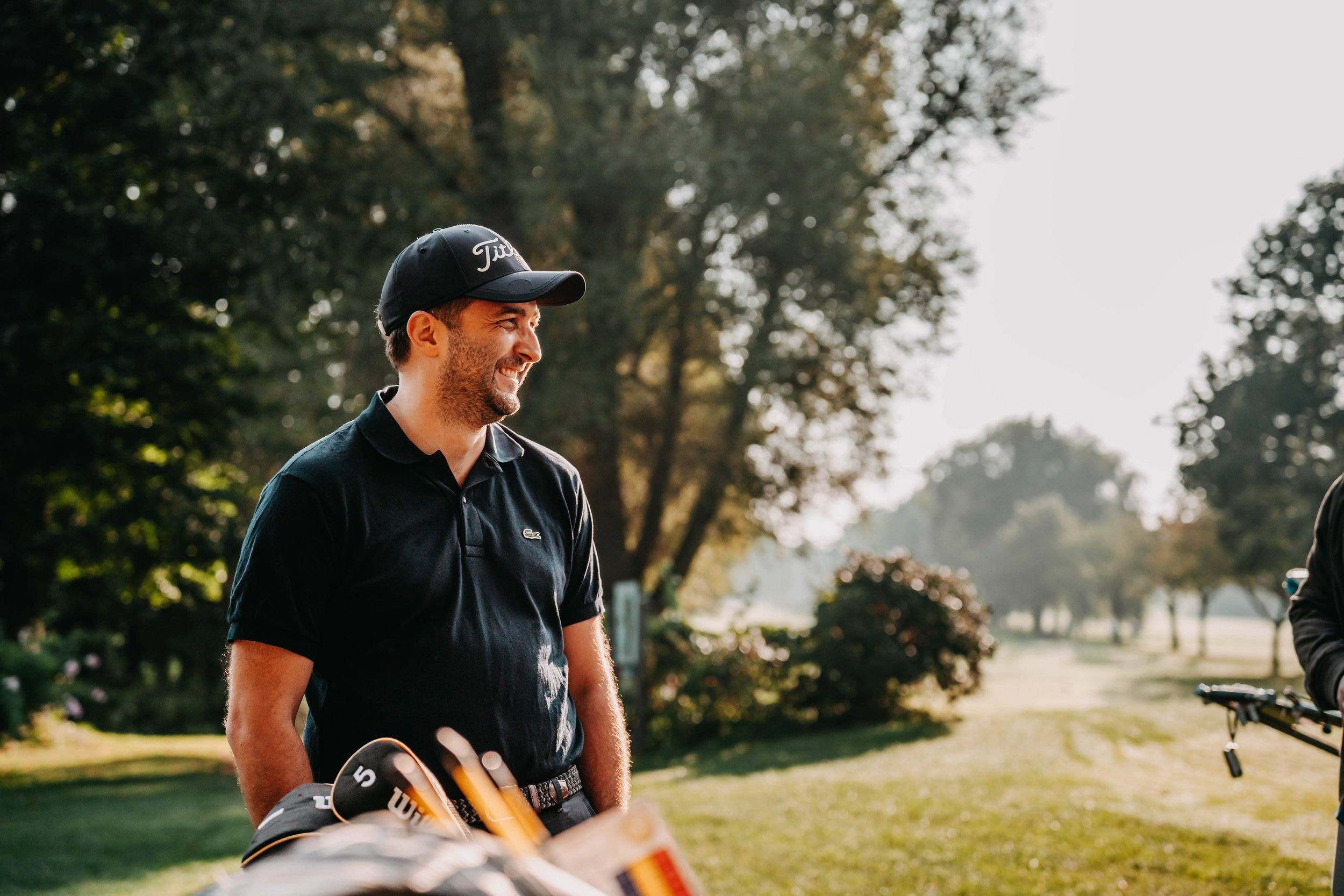 Lächelnder Mann mit Kappe und schwarzem Poloshirt auf dem Golfplatz im Gegenlicht.