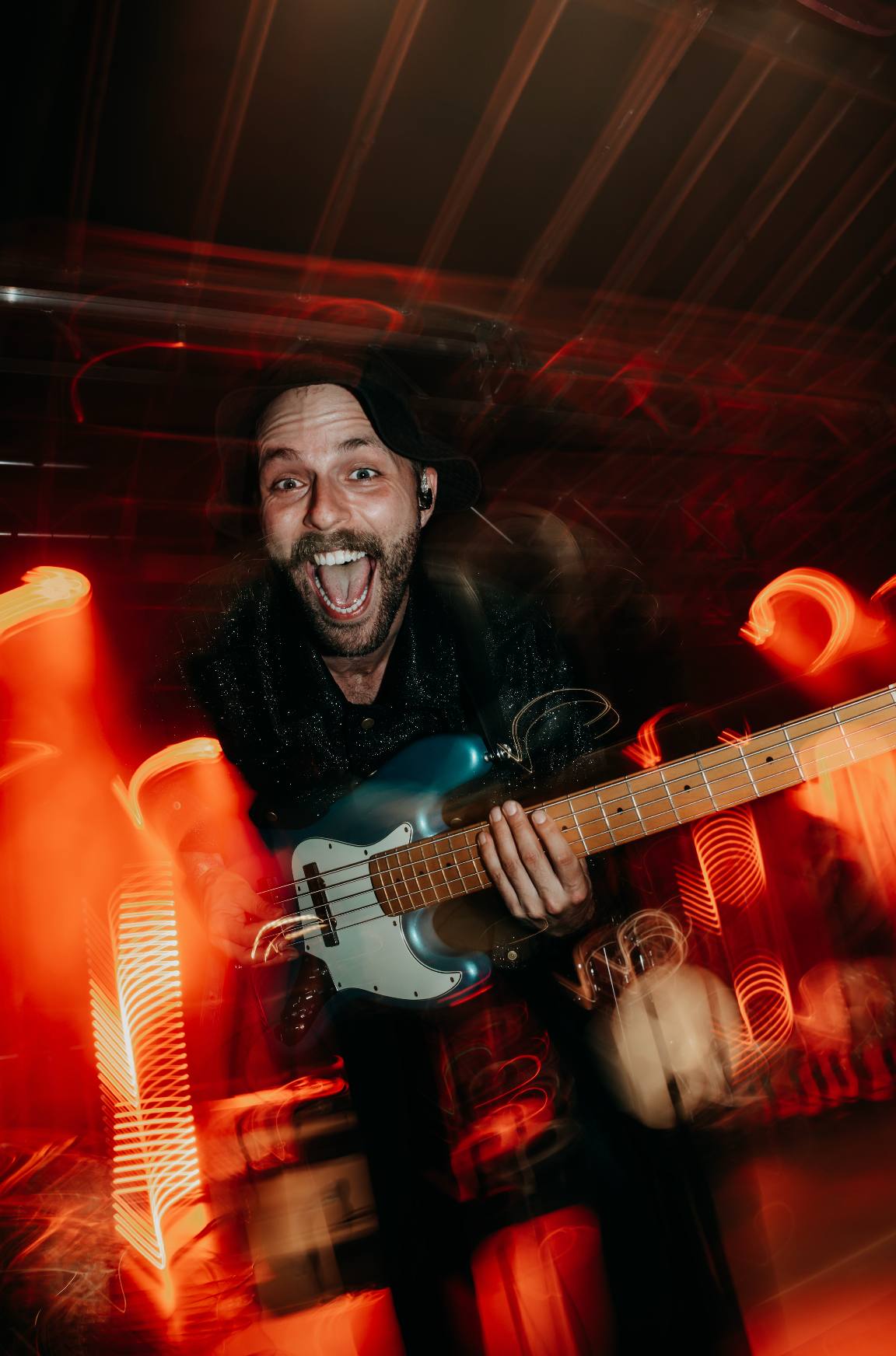 Lachender Bassist auf der Bühne beim Sommerfest, umgeben von roten und orangefarbenen Lichtspuren.