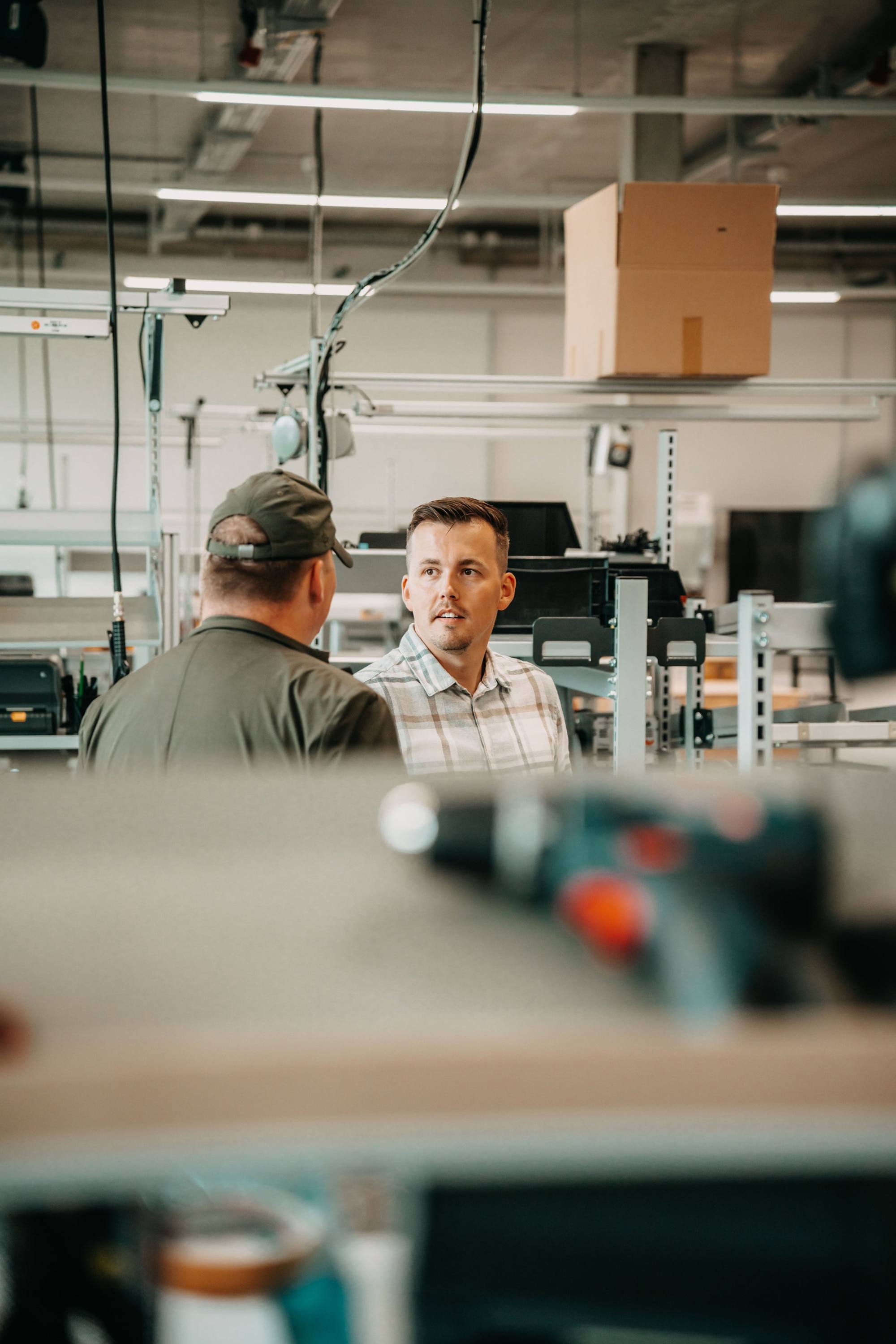 Zwei Männer reden in einer Werkstatt oder Fabrikhalle, umgeben von technischen Geräten und Regalen.