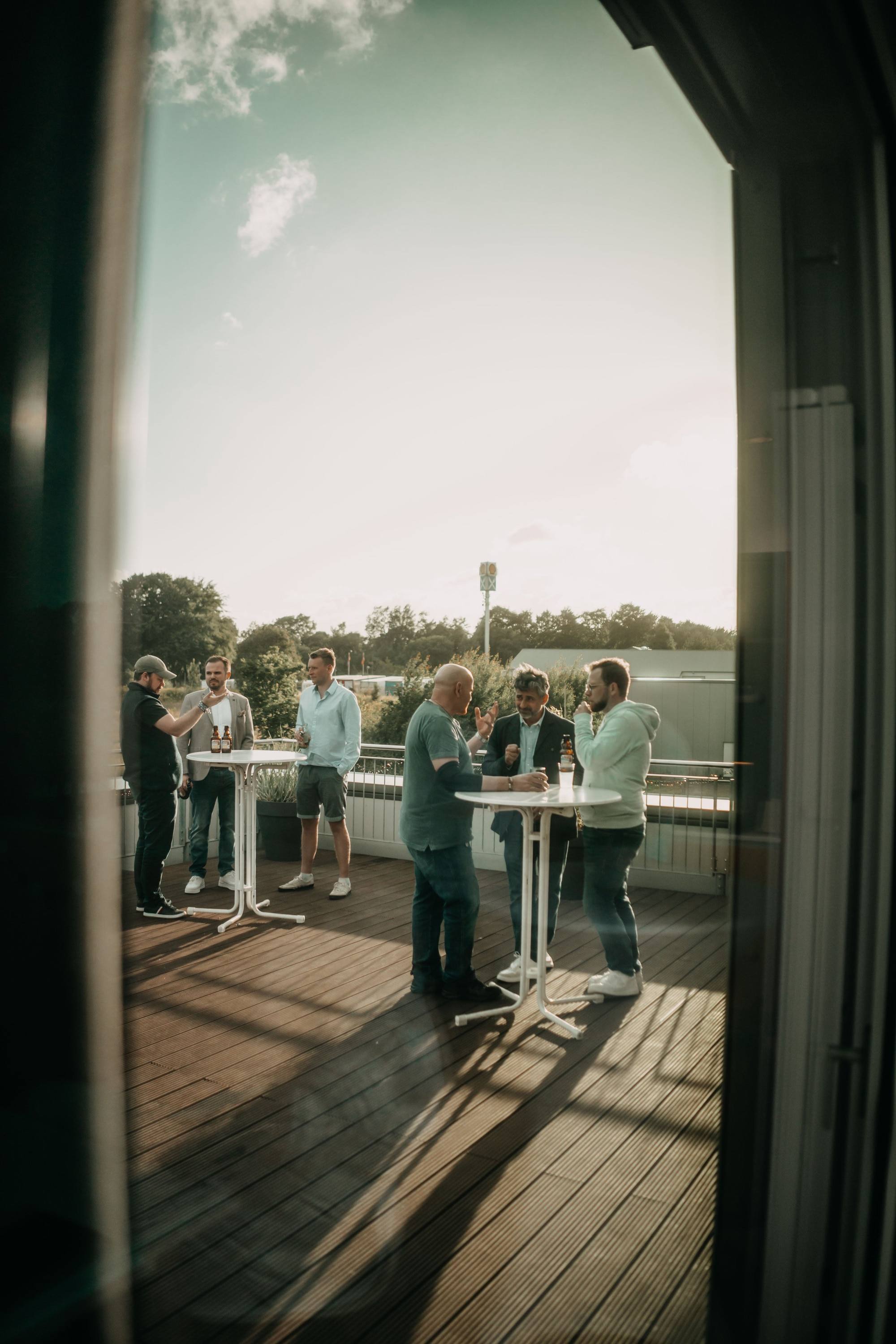 Menschen auf einer Dachterrasse beim geselligen Beisammensein bei Sonnenuntergang, entspannte Atmosphäre, sechs Männer in kleinen Gruppen, sprechen und trinken Bier.