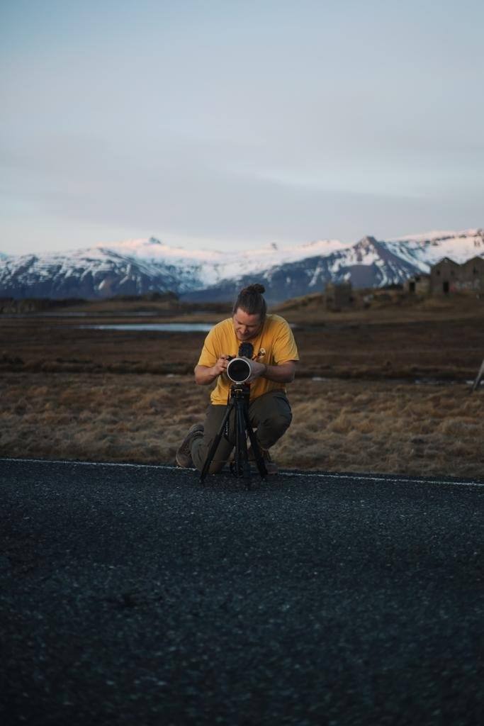 Eine Person in gelbem T-Shirt fotografiert mit einer Kamera auf einem Stativ auf einer Straße vor einer Landschaft mit Bergen im Hintergrund.