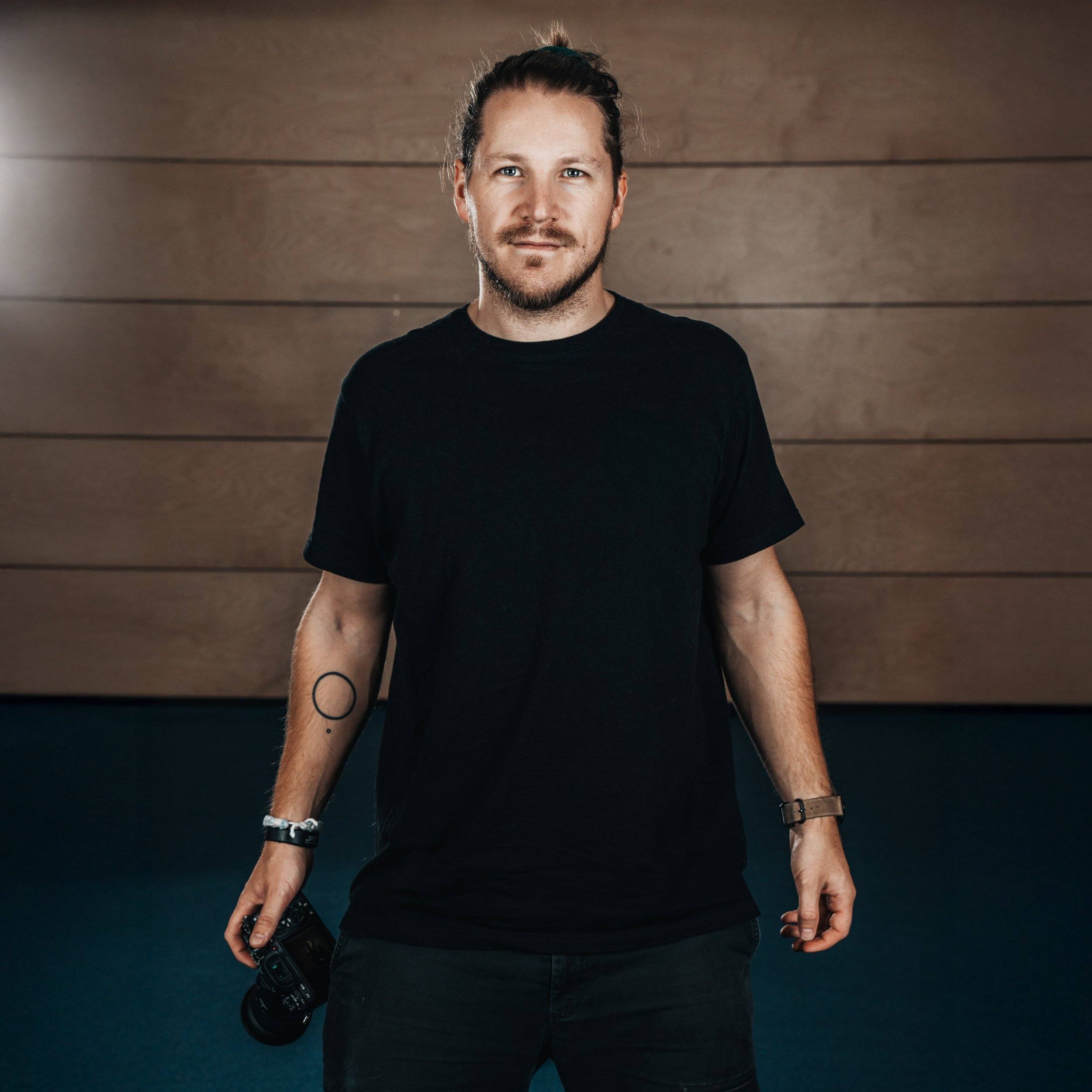 Junge Mann mit schwarzen T-Shirt, im Hintergrund eine Holzvertäfelung, hält eine Kamera in der rechten Hand, tragen eine Armbanduhr und ein Tattoo am linken Arm.